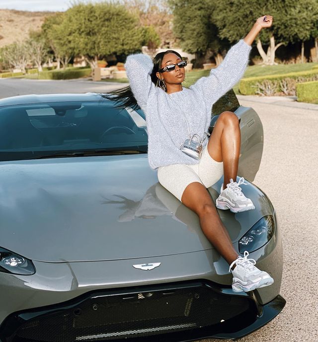 Itali Miller in a grey sweat shirt and white shorts posing on her car.
